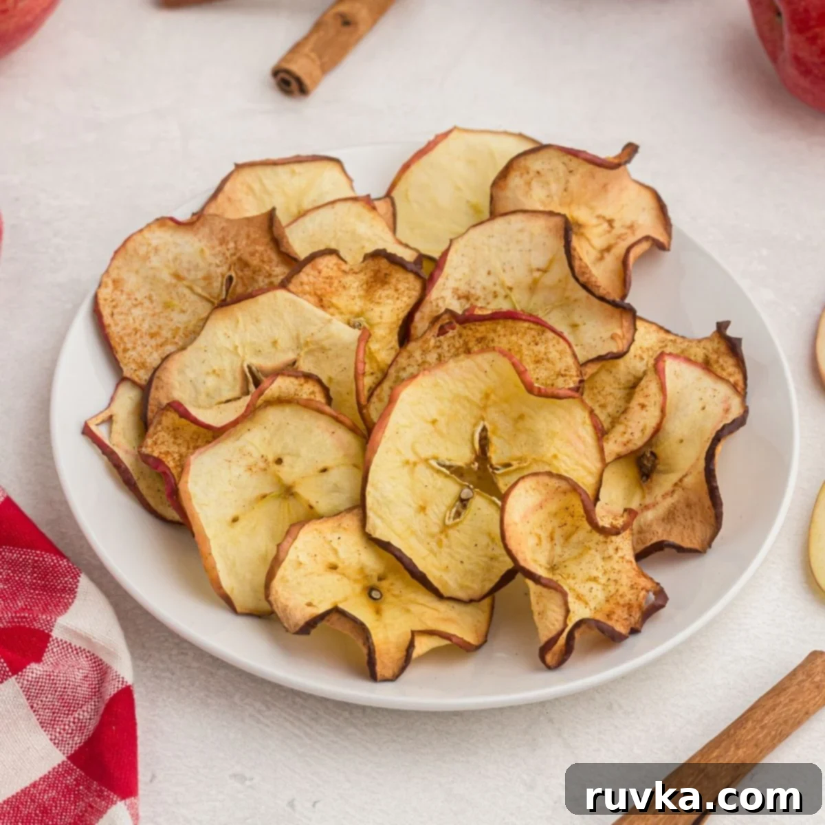 Effortless Holiday Feasts: 15 Air Fryer Recipes 11 golden apple slices, cooked into chips. Served on a white plate with a red napkin and cinnamon sticks on the table.