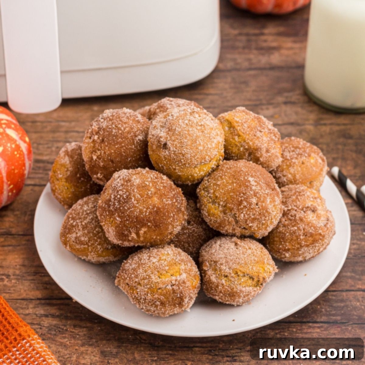 Effortless Holiday Feasts: 15 Air Fryer Recipes 16 Golden pumpkin donut holes stacked on a white plate in front of glasses of milk and air fryer on a wooden table.