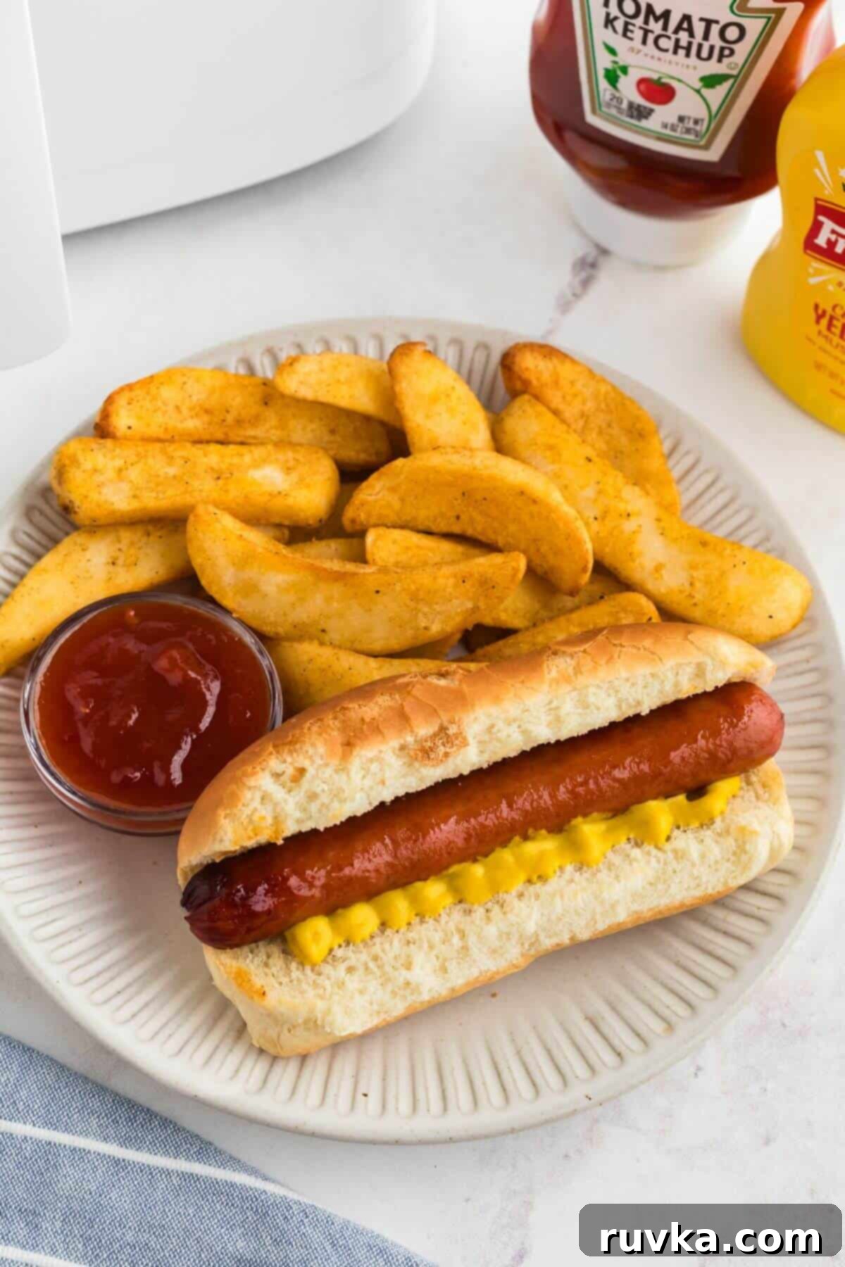 Easy Air Fryer Frozen Hot Dogs 2 Juicy cooked hot dog in a bun with French fries on a white plate.