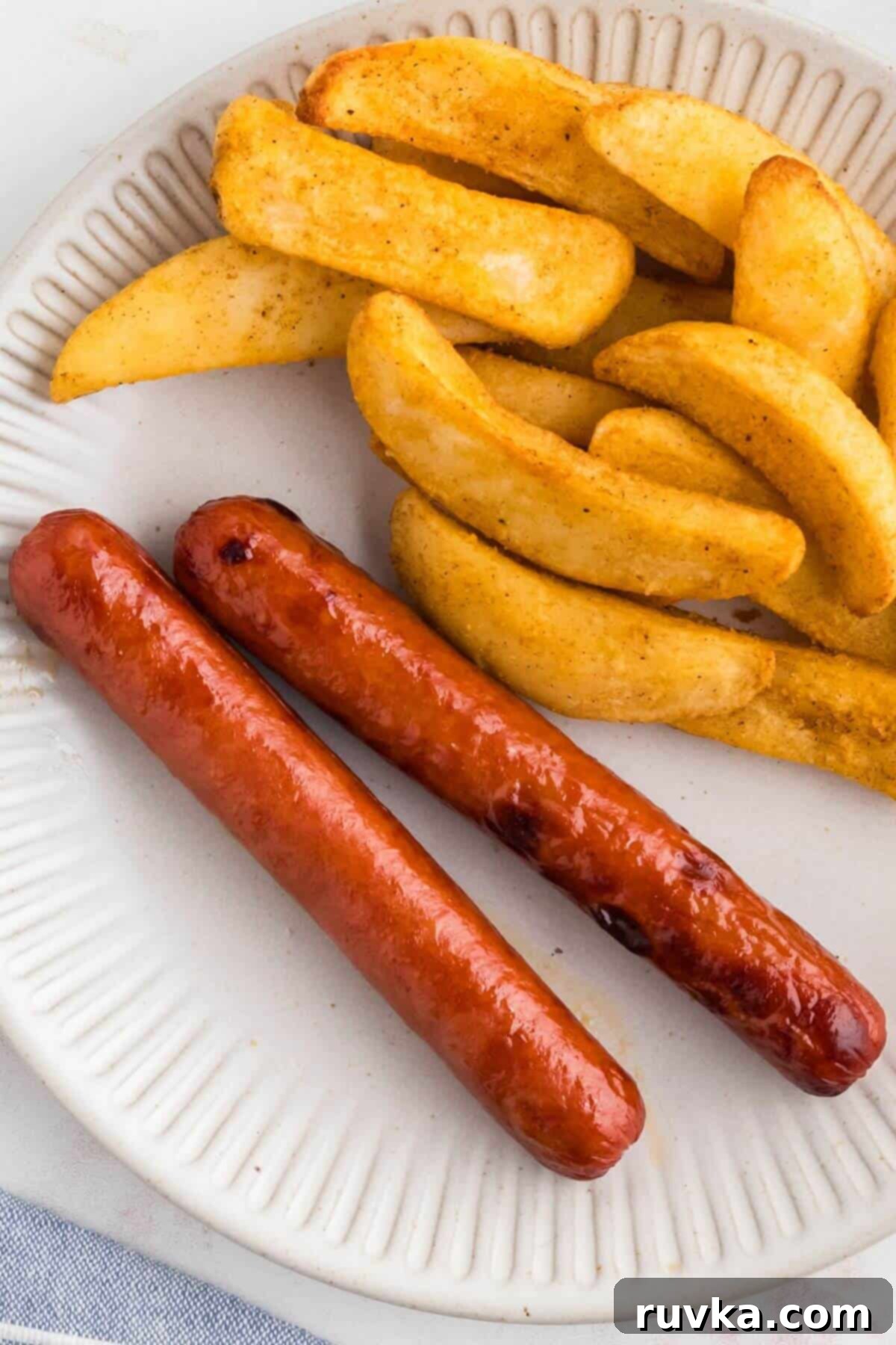 Easy Air Fryer Frozen Hot Dogs 6 Juicy cooked hot dogs on a white plate with crispy french fries as a side dish.