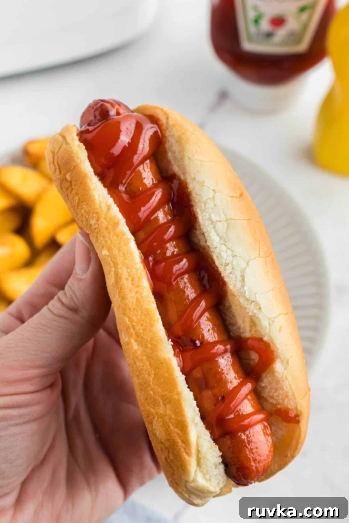 Easy Air Fryer Frozen Hot Dogs 7 Juicy golden brown hot dog in a bun with ketchup drizzled on top being held over a plate.