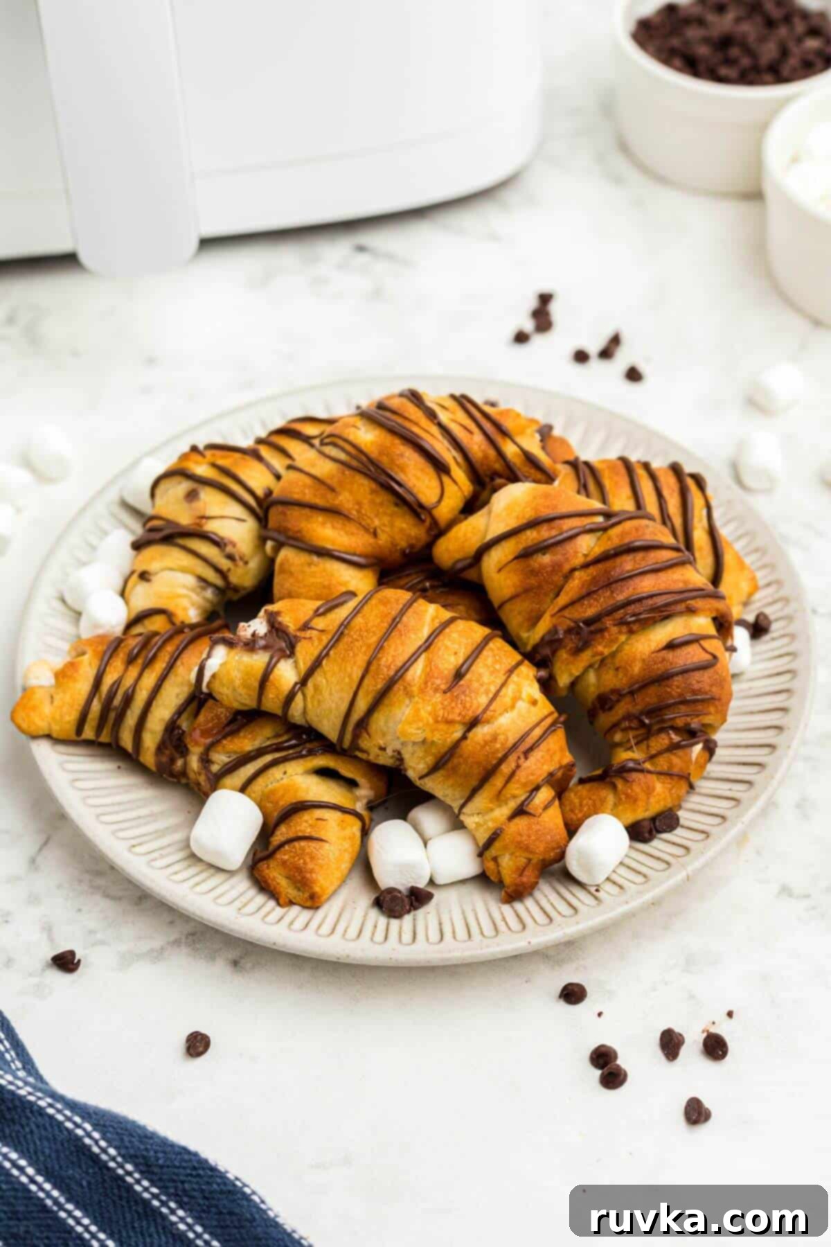 Air Fryer S'mores Crescent Rolls 2 Golden flaky crescent rolls filled with marshmallows and chocolate chips, stacked on a cream plate with a blue linen.