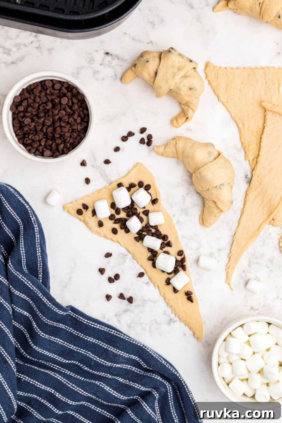 Air Fryer S'mores Crescent Rolls 4 Unrolled crescent rolls topped with mini chocolate chips and mini marshmallows on a marble table being rolled into crescent shapes.