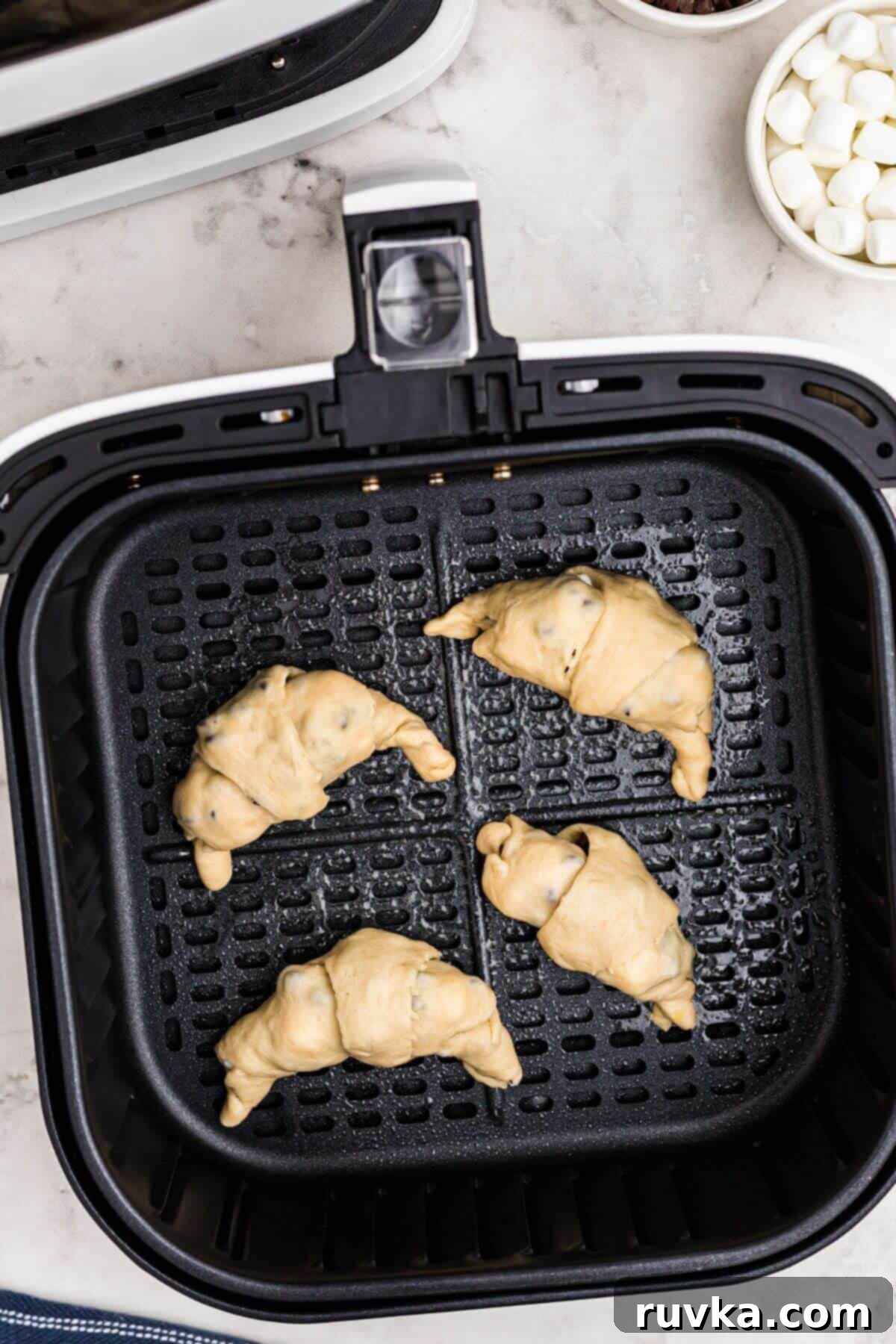 Air Fryer S'mores Crescent Rolls 5 Uncooked rolled crescent rolls filled with mini chocolate chips and mini marshmallows, in an air fryer basket.