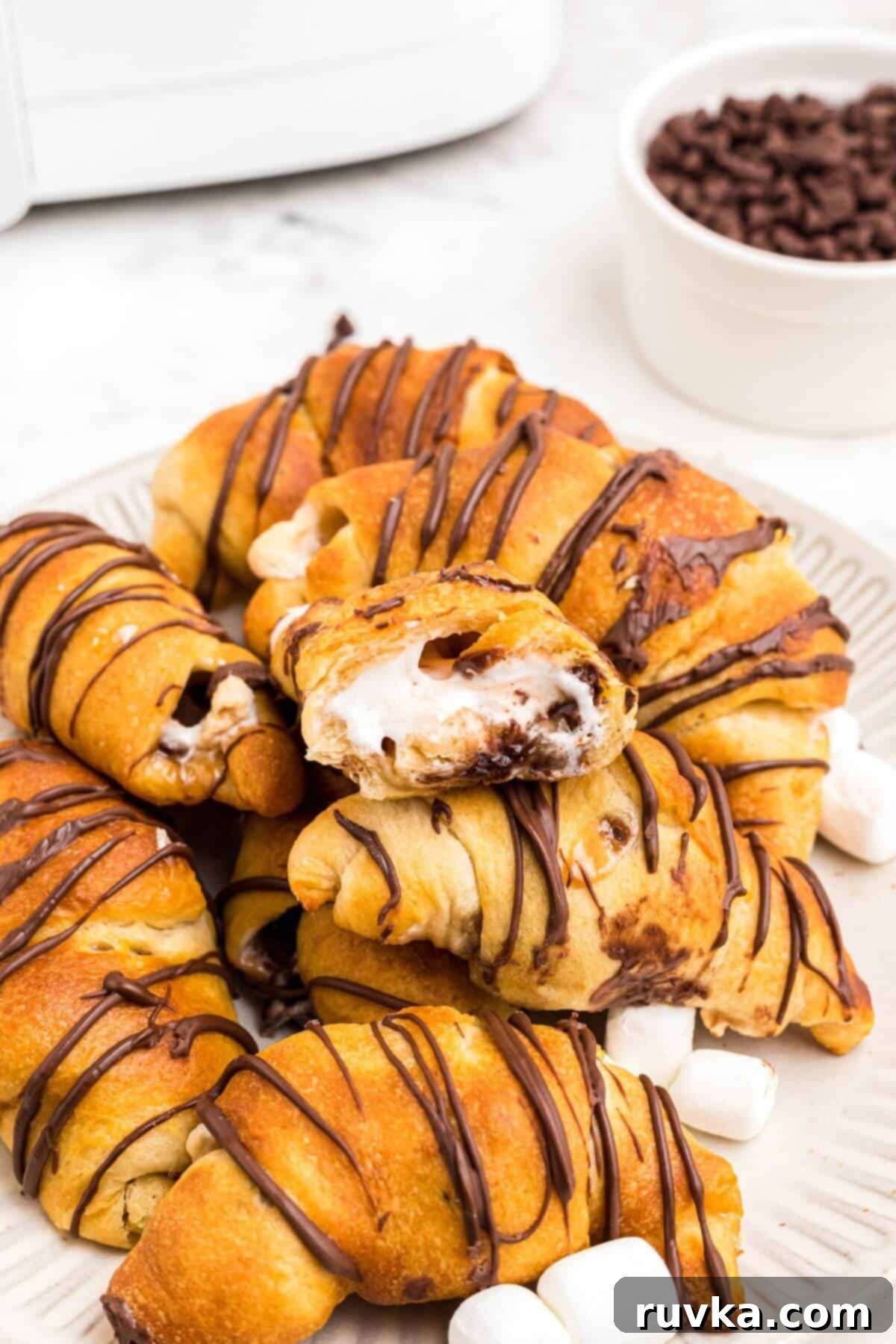 Air Fryer S'mores Crescent Rolls 7 Golden flaky crescent rolls filled with gooey marshmallows and melty chocolate chips stacked on a plate with mini marshmallows and chips on the plate.