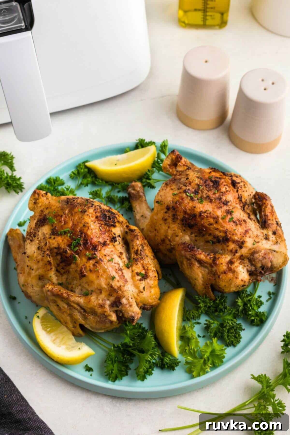 Crispy Air Fryer Cornish Game Hen 8 Golden brown crispy Cornish hens on a light blue plate with lemon wedges and parsley garnish on a white marble table.