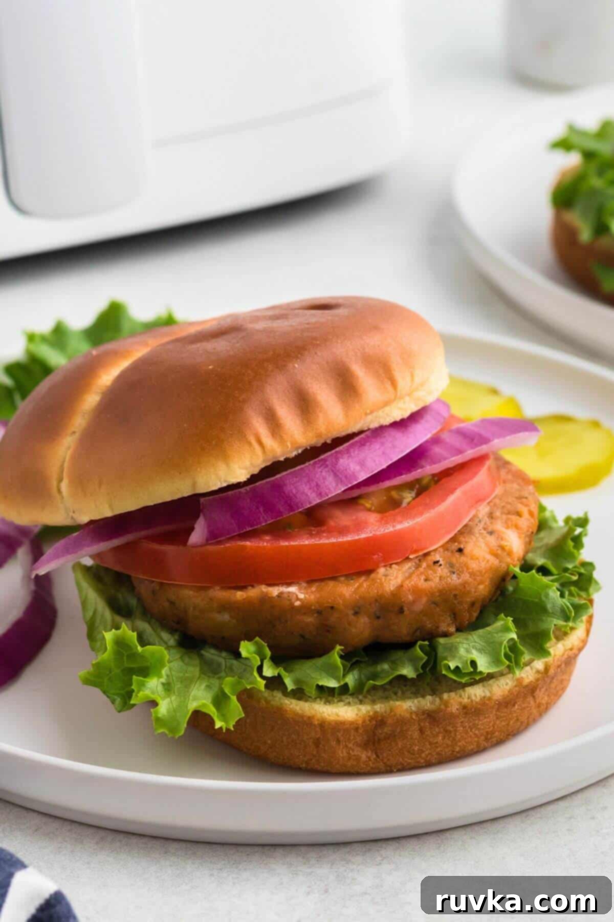 Crispy Air Fryer Frozen Salmon Burgers 2 Salmon burger on a bun over lettuce, then topped with slice of tomato and red onion on a white plate.