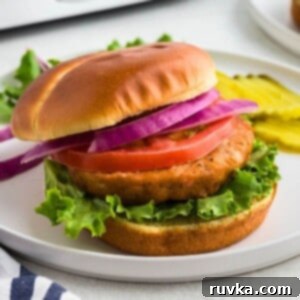 Crispy Air Fryer Frozen Salmon Burgers 7 Salmon burger on a bun with lettuce, tomato, and sliced onion on a white plate.