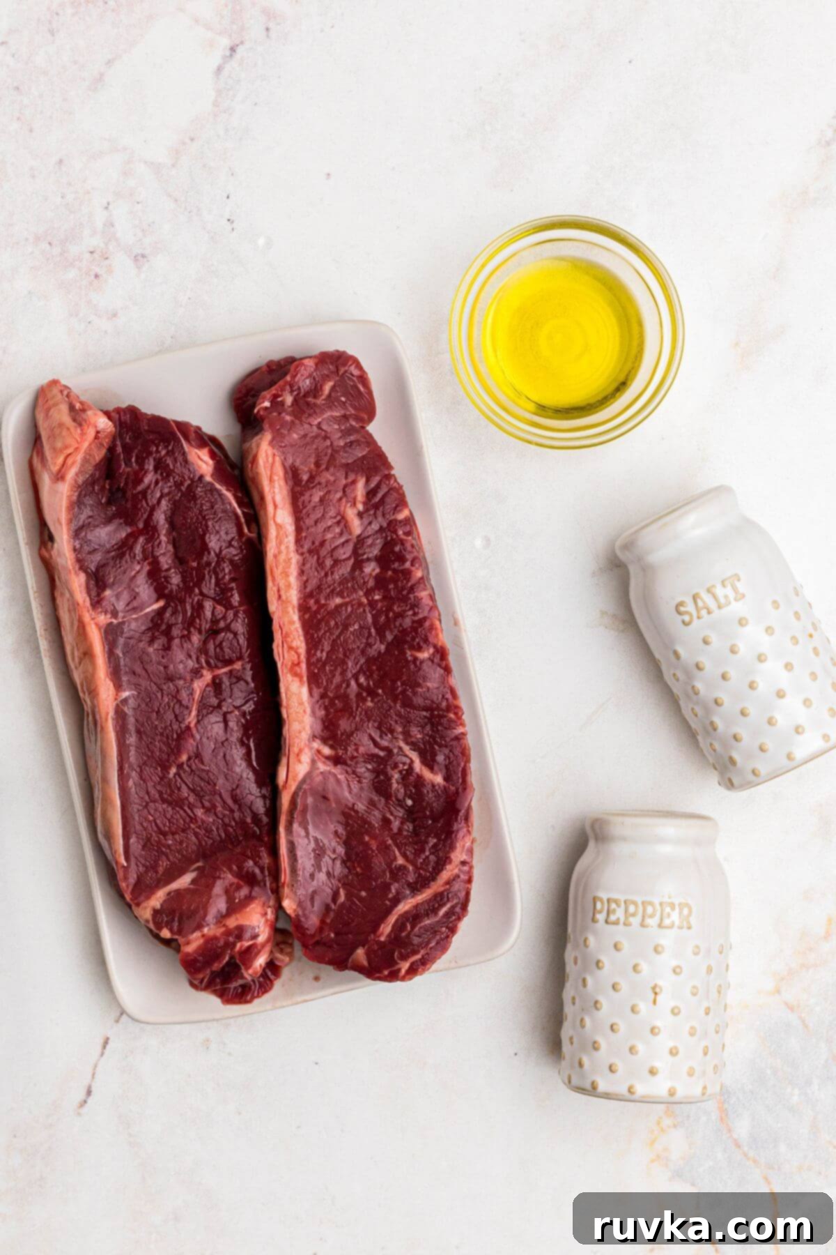 Ingredients for Air Fryer New York Strip Steak Uncooked New York strip steaks on a marble surface, seasoned with olive oil, salt, and pepper, ready for the air fryer.