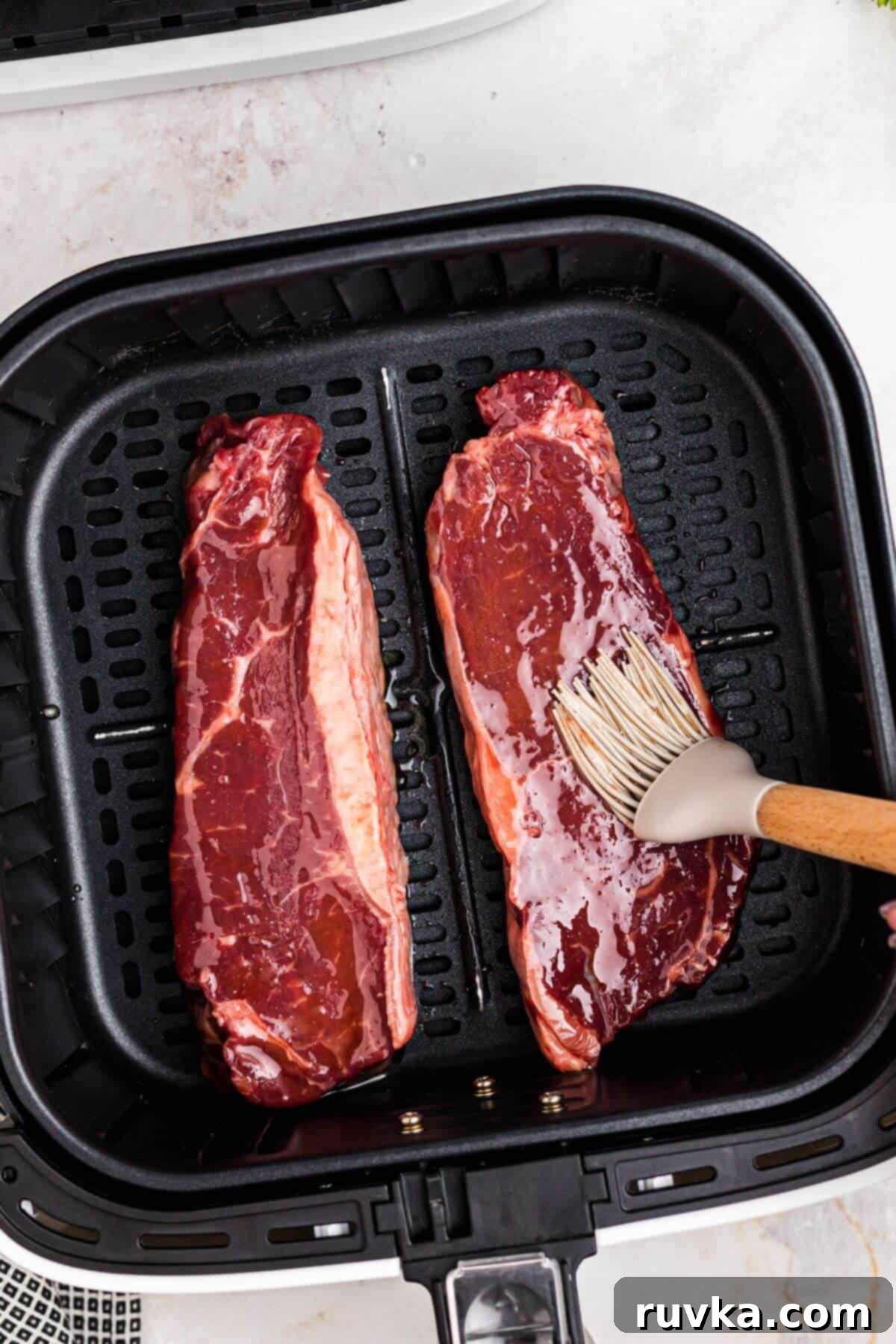 Seasoning New York Strip Steak for Air Frying Uncooked New York strip steaks being brushed with olive oil in the air fryer basket.