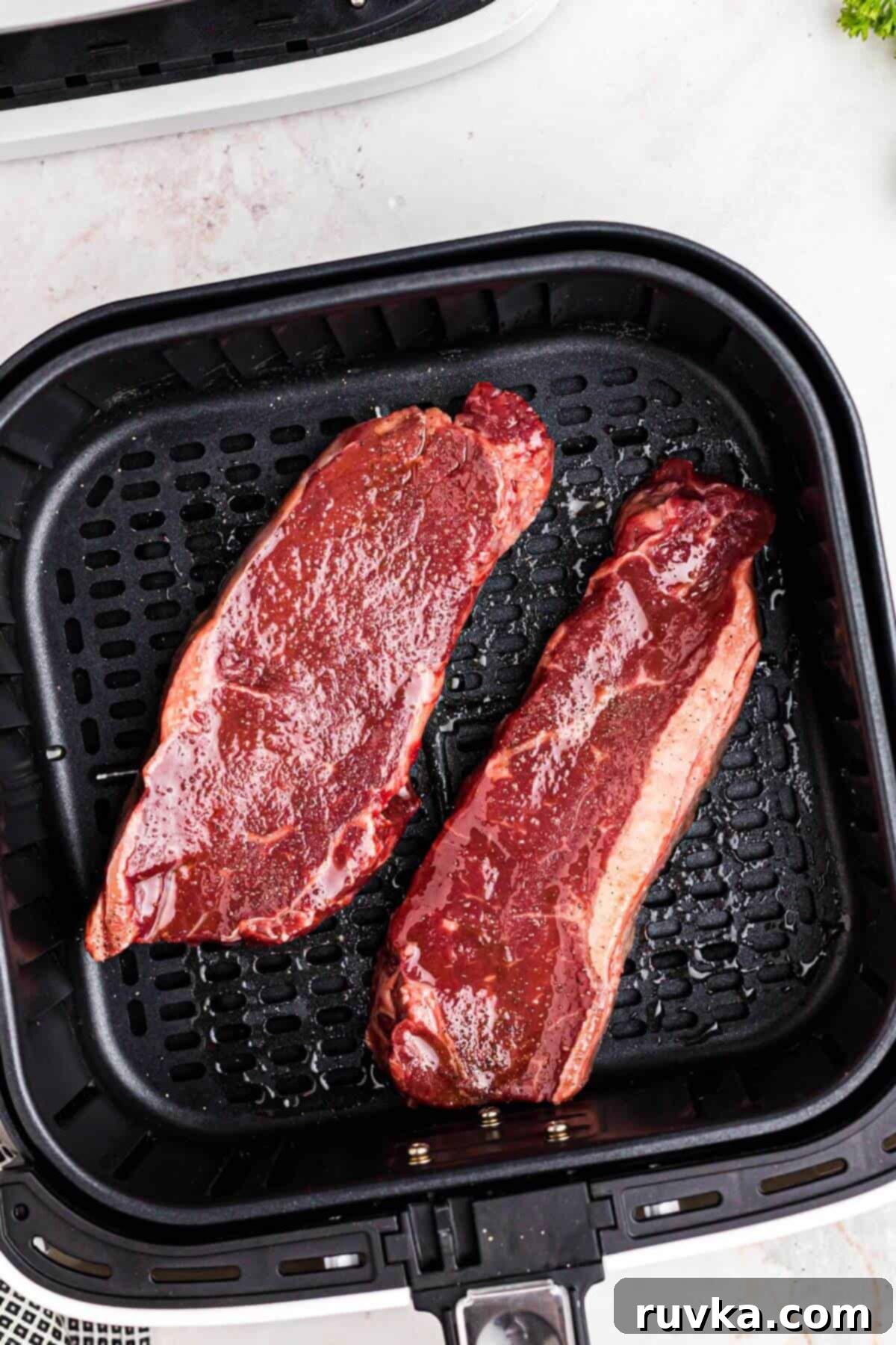 Steaks in Air Fryer Basket Seasoned uncooked New York strip steaks neatly arranged in a single layer within the air fryer basket.