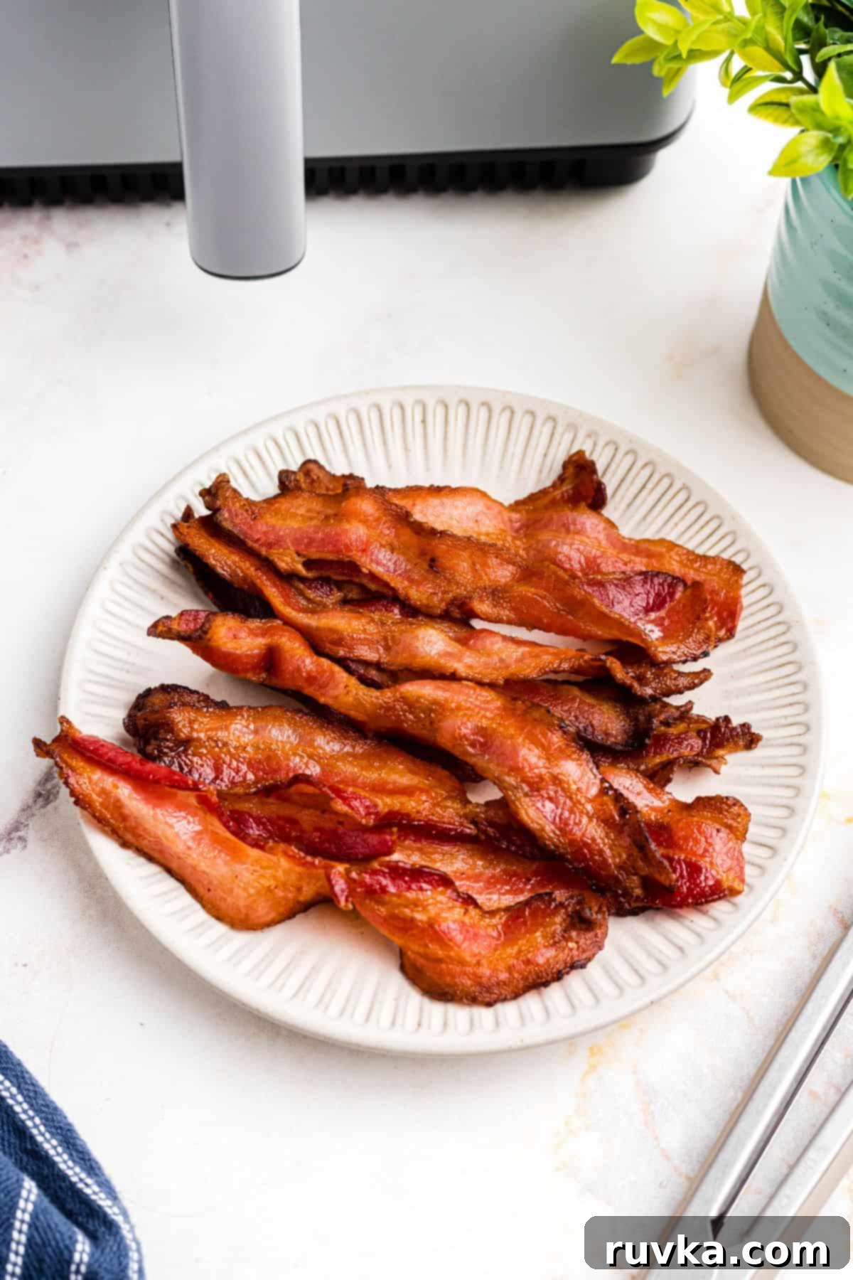 Air Fryer Frozen Bacon Hack 2 Golden crispy bacon stacked on a cream colored plate in front of a gray air fryer.
