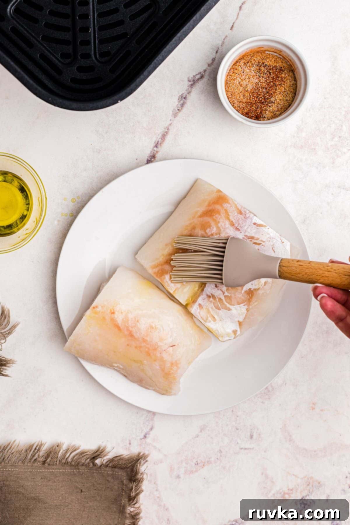 Perfect Air Fryer Frozen Cod 4 Brushing olive oil on frozen cod filets on a white plate before air frying.