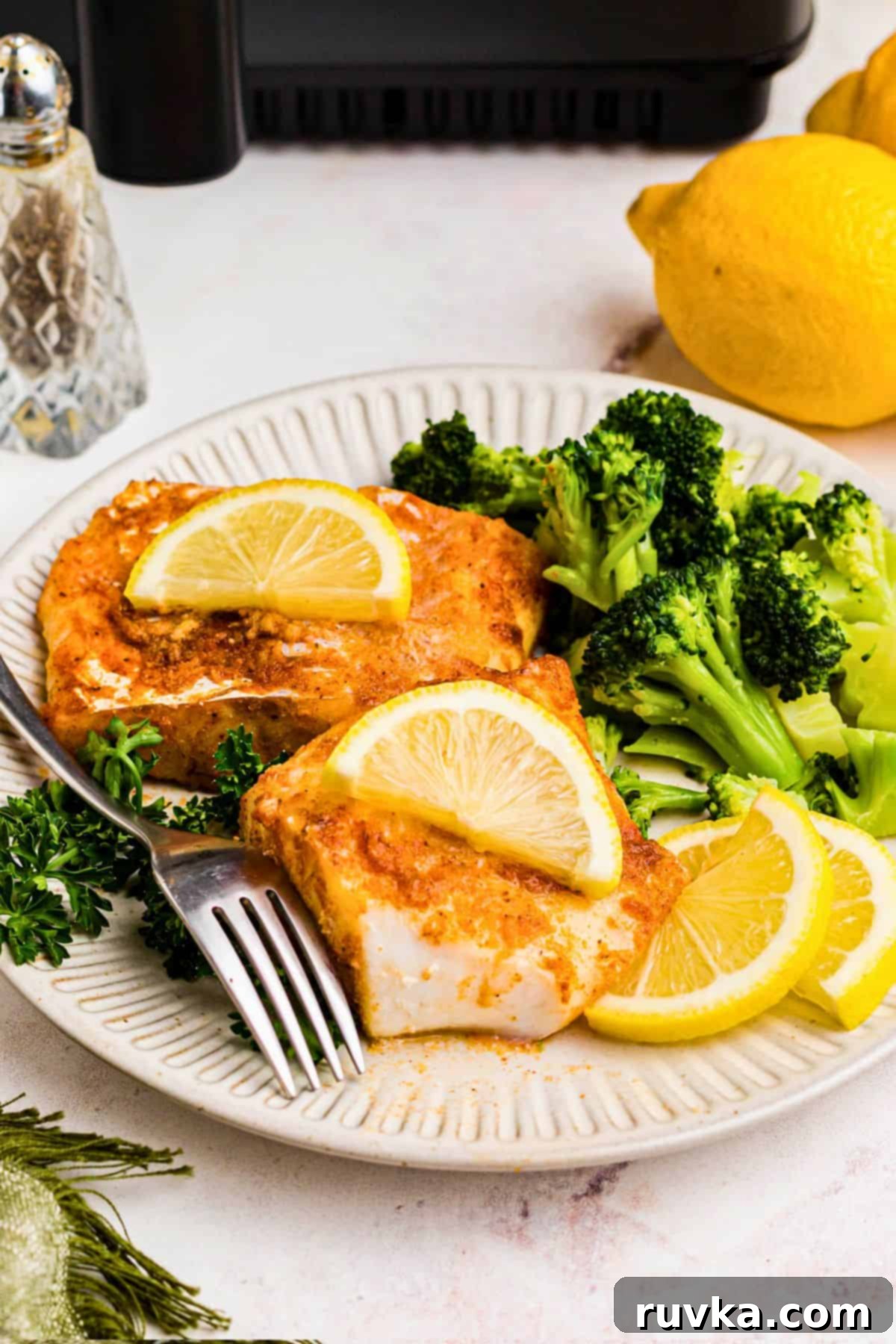 Perfect Air Fryer Frozen Cod 8 Cooked and seasoned cod on a white plate with broccoli and lemon slices, with a fork laying across the plate.