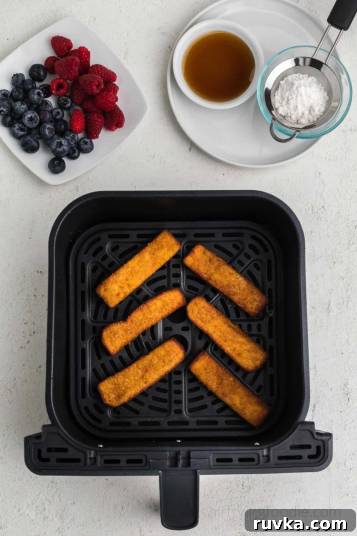 Crispy Air Fryer Frozen French Toast Sticks 5 french toast sticks in air fryer basket, fully cooked from frozen. Berries, syrup, and powdered sugar above the basket.