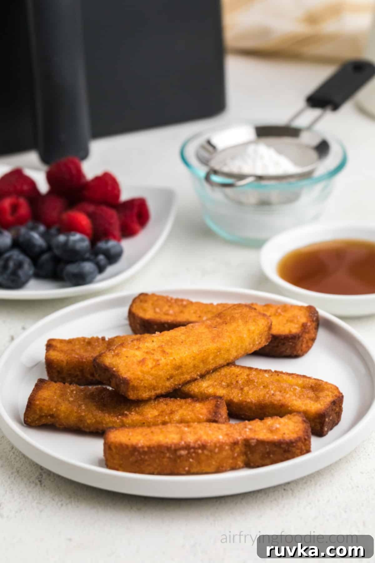 Crispy Air Fryer Frozen French Toast Sticks 6 Air fried frozen french toast sticks, fully cooked and served on a white plate with berries, syrup, and powdered sugar in the background.