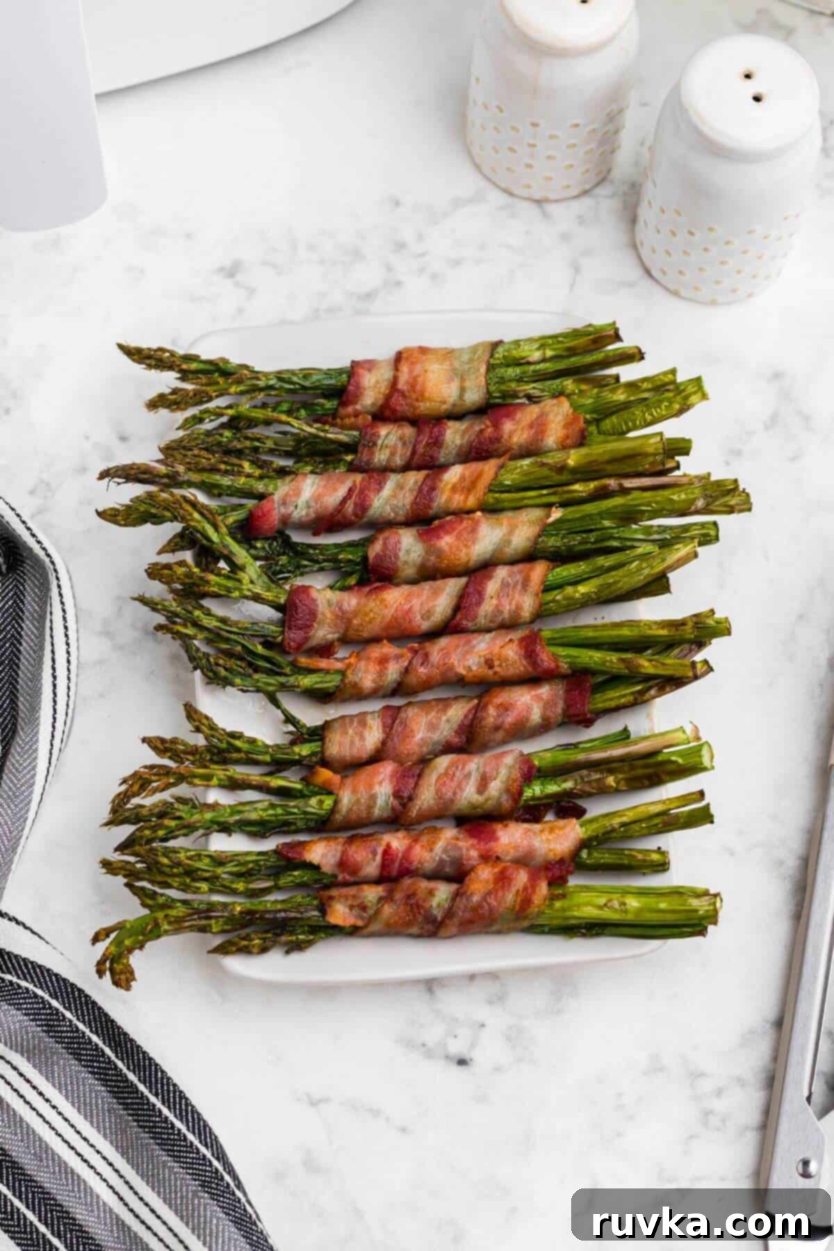 Crispy Air Fryer Bacon Wrapped Asparagus 2 Crispy brown bacon wrapped around asparagus spears, on a white rectangle plate.