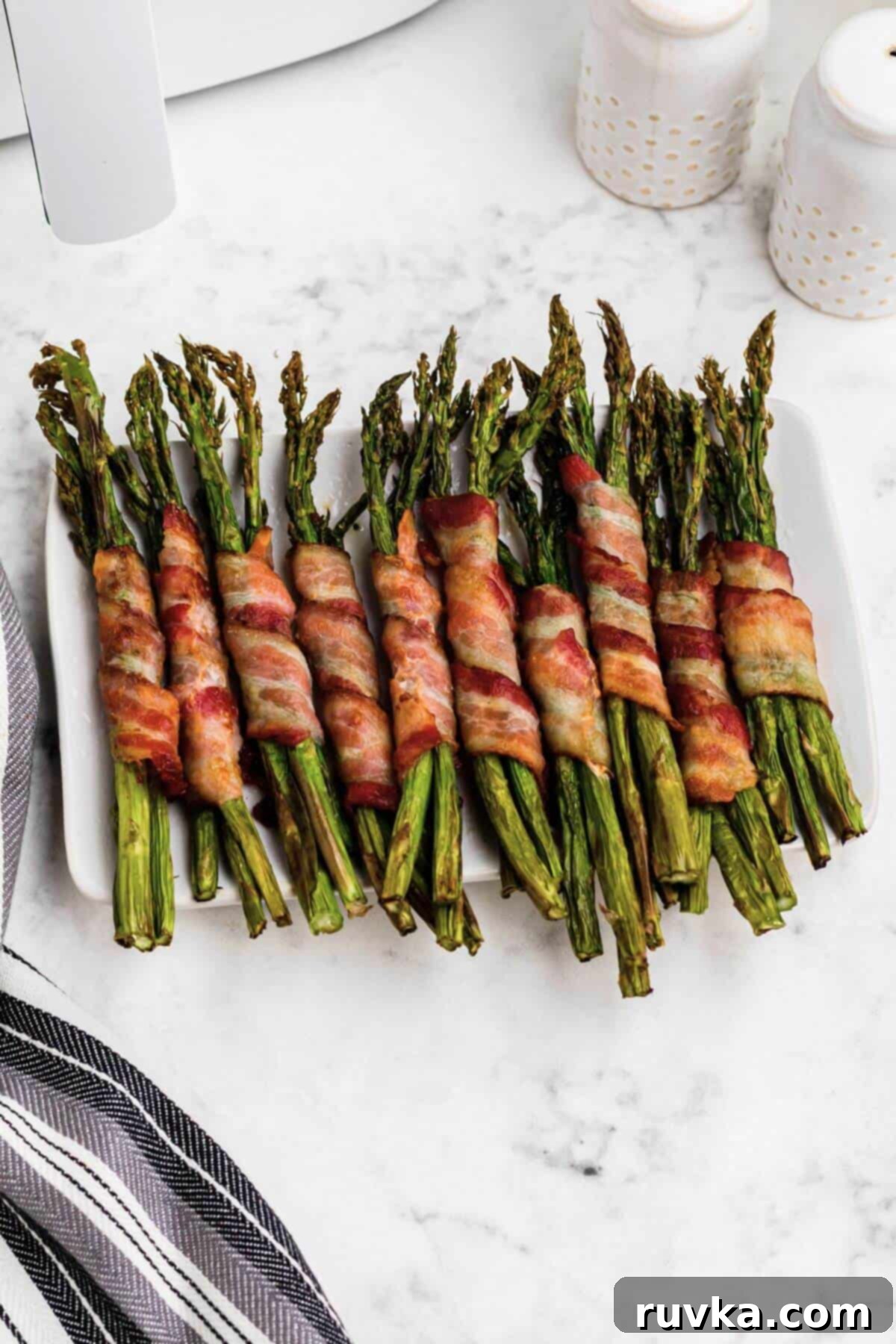 Crispy Air Fryer Bacon Wrapped Asparagus 6 Crispy golden brown bacon wrapped around asparagus stalks on a white rectangle plate.