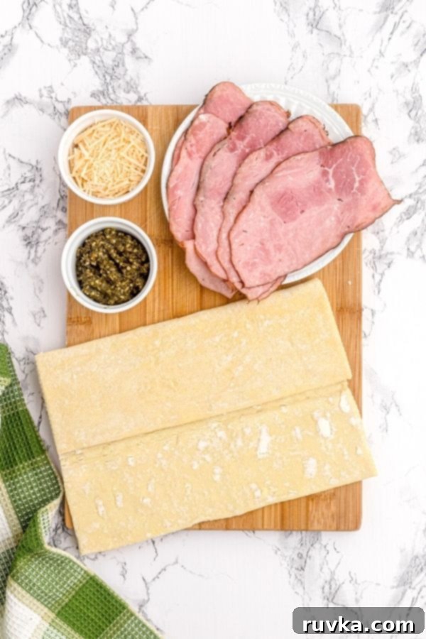 Crispy Air Fryer Pesto Pinwheels 3 Ingredients needed to make pesto pinwheels on a cutting board including frozen puff pastry, a jar of basil pesto, ham slices, and a bowl of shredded Parmesan cheese.