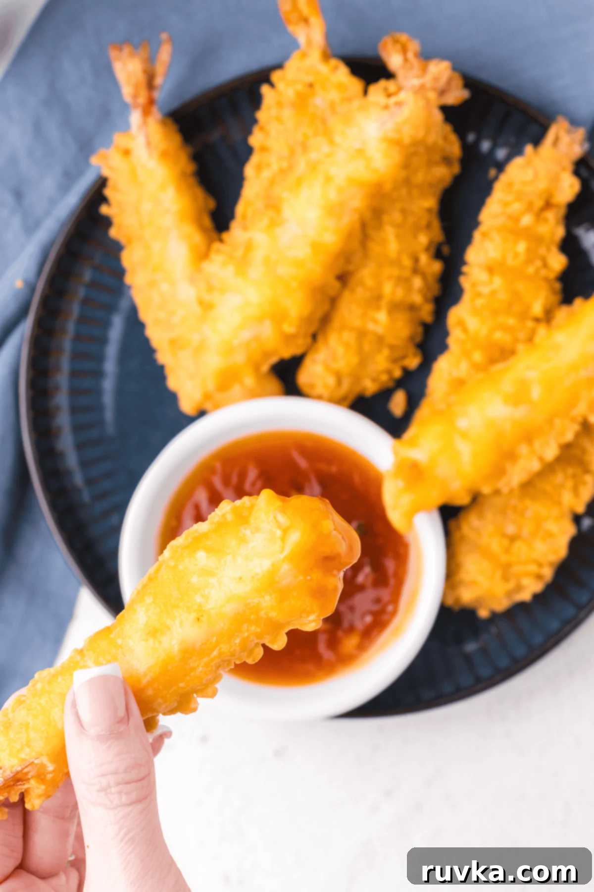Crispy Air Fryer Frozen Shrimp Tempura 7 A hand holding a piece of shrimp tempura, dipping it into a small bowl of sauce, with more tempura in the background.