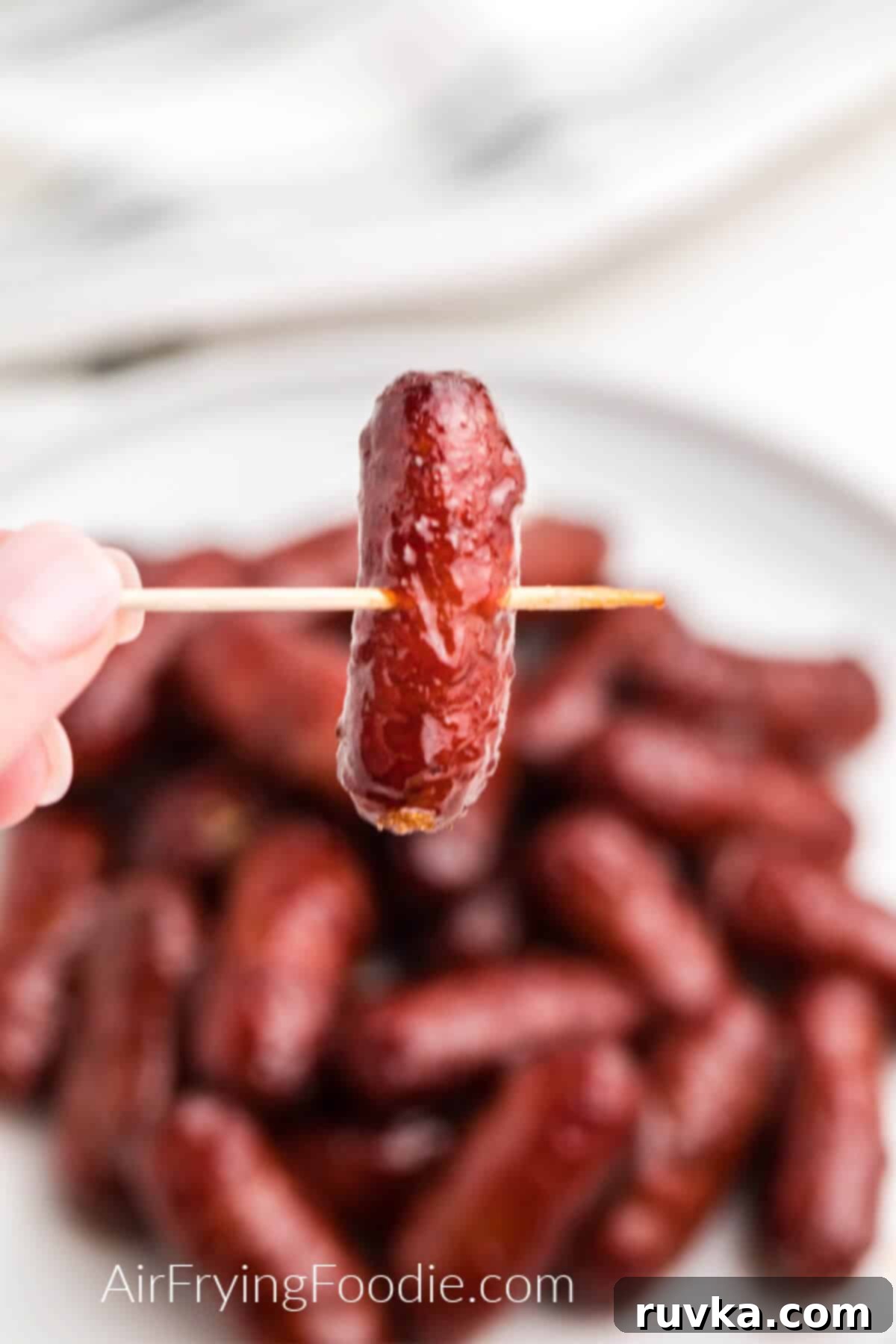 Easy Air Fryer BBQ Little Smokies 6 BBQ Little Smokie on a toothpick ready to eat with a plate full of cooked little smokies in the background.