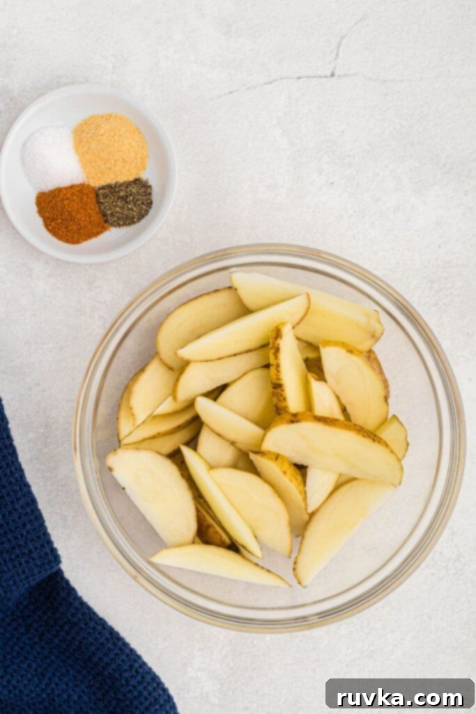 Crispy Air Fryer Potato Wedges 3 Uncooked potato wedges in a clear glass bowl with seasonings in a small dish on a marble table.