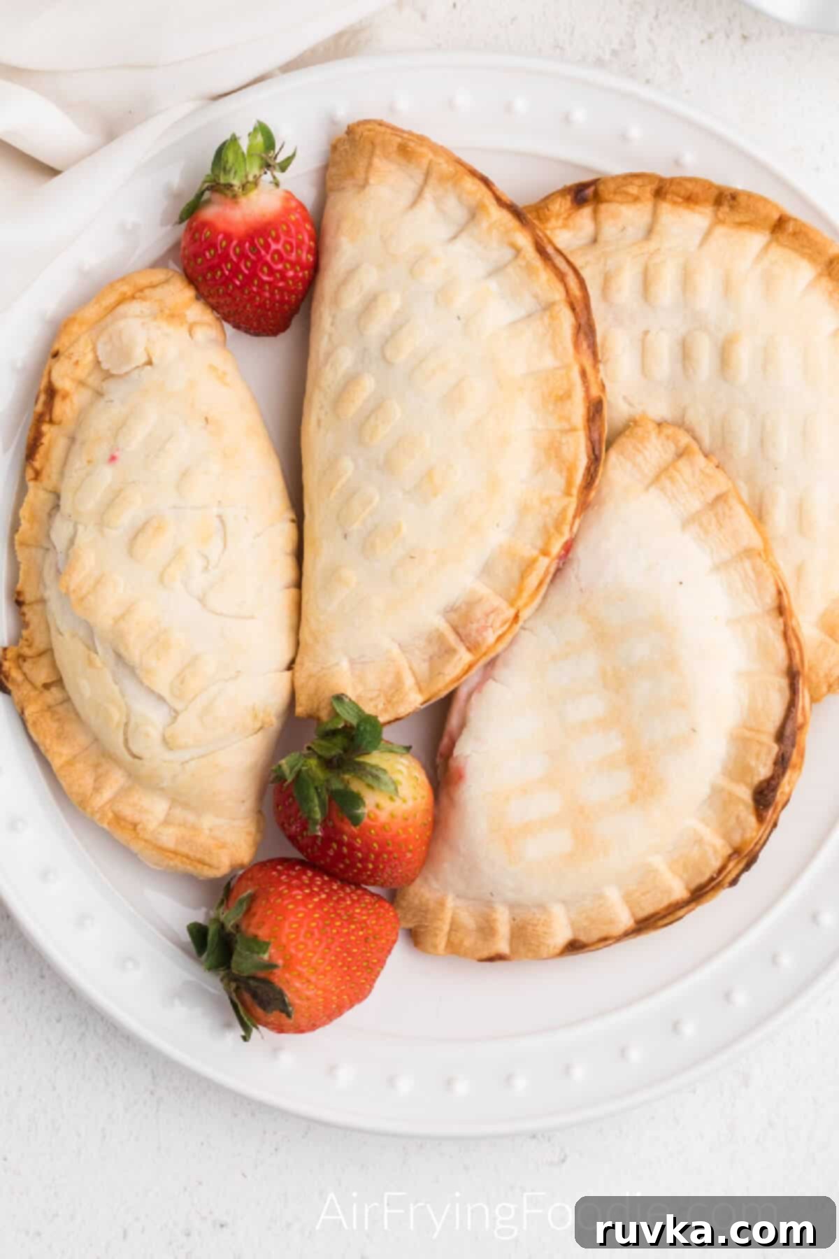 Easy Air Fryer Strawberry Cream Cheese Pie 2 Air fried strawberry cream cheese hand pies on a white plate with fresh strawberries, ready to serve.