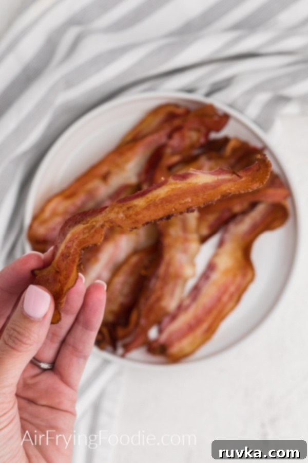 Crispy Air Fryer Bacon 2 Hand holding crispy bacon over a plate of bacon that was made in the air fryer.