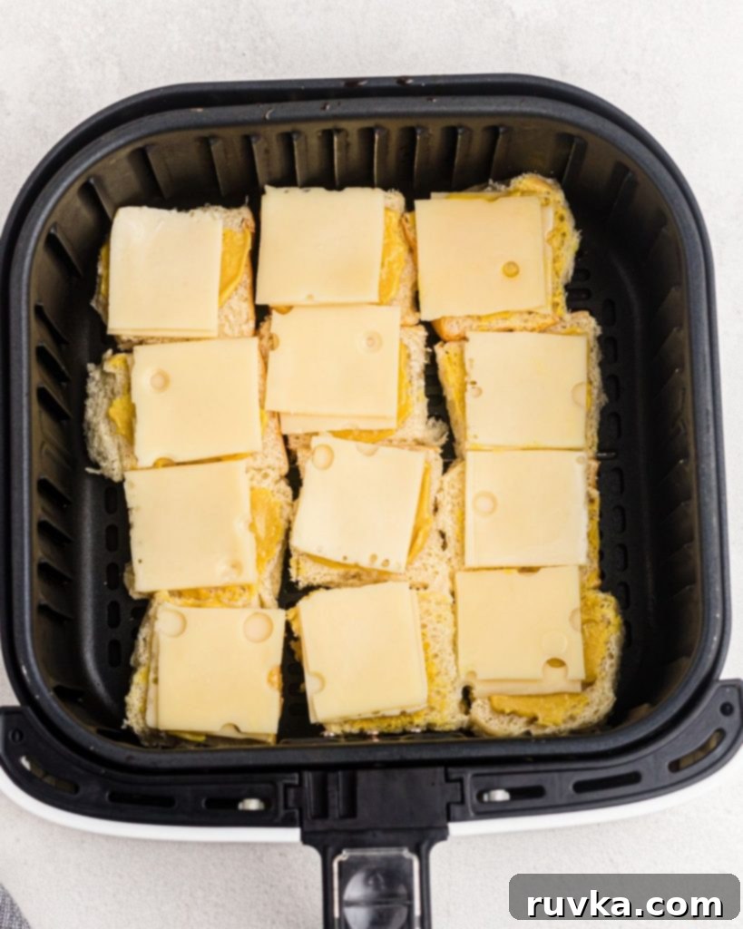 Irresistible Air Fryer Ham and Cheese Sliders 5 The bottom halves of Hawaiian rolls neatly arranged in an air fryer basket, topped with honey mustard and folded cheese slices, ready for the ham.