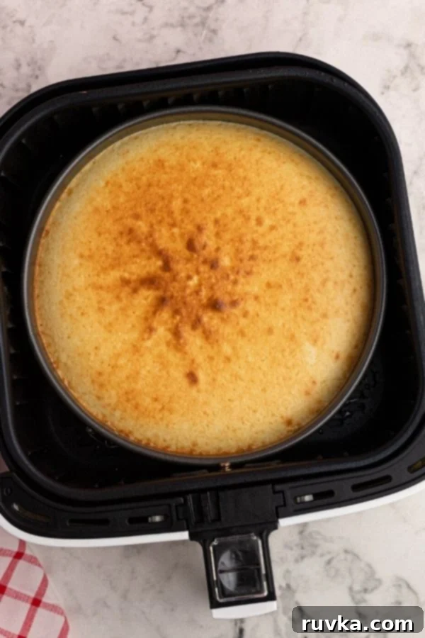 Effortless Air Fryer Cheesecake 4 Cooked cheesecake in a pan, carefully placed inside the basket of an air fryer after baking, with a lightly golden top.