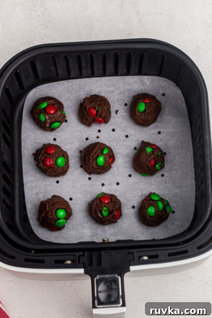Quick Christmas Air Fryer Brownie Bites 6 Neatly scooped balls of festive brownie cookie dough, studded with colorful candies, precisely arranged on parchment paper within the air fryer basket, prepped and ready for baking.