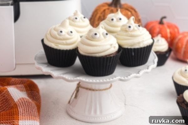 Spooktacular Air Fryer Ghost Cupcakes 2 Ghost cupcakes on a white stand in front of an air fryer with pumpkins in the background.