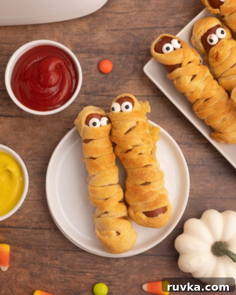 Spooky Air Fryer Hot Dog Mummies 3 A plate of golden-brown Air Fryer Mummy Dogs, artfully arranged with mustard and ketchup for dipping, surrounded by scattered Halloween candies, setting a festive mood.