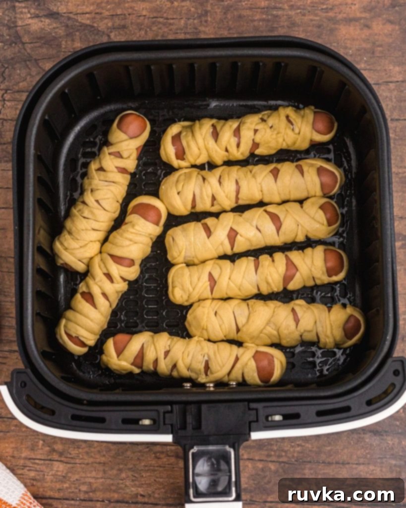 Spooky Air Fryer Hot Dog Mummies 6 Several hot dogs are wrapped with crescent roll strips, arranged to resemble mummies, and placed neatly in an air fryer basket, ready for cooking.