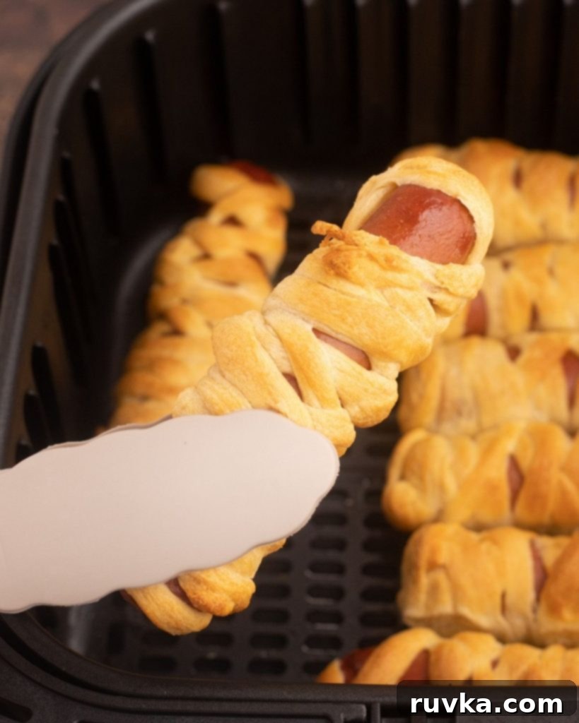 Spooky Air Fryer Hot Dog Mummies 8 A single cooked Air Fryer Mummy Dog, perfectly golden and wrapped in crescent dough, being lifted out of the air fryer basket with tongs, ready for its candy eyes and serving.