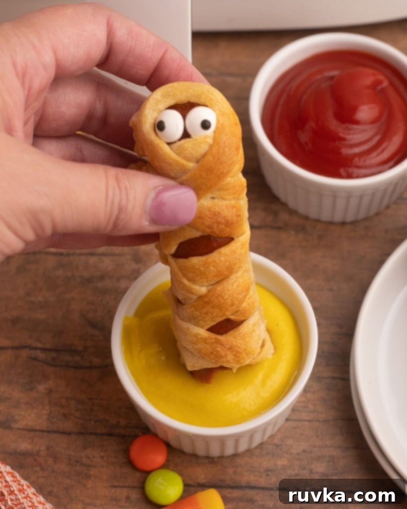 Spooky Air Fryer Hot Dog Mummies 9 A close-up shot of a golden-brown Air Fryer Mummy Dog, held by tongs, being dipped into a small bowl of mustard, with festive Halloween candies scattered on a wooden table in the background.