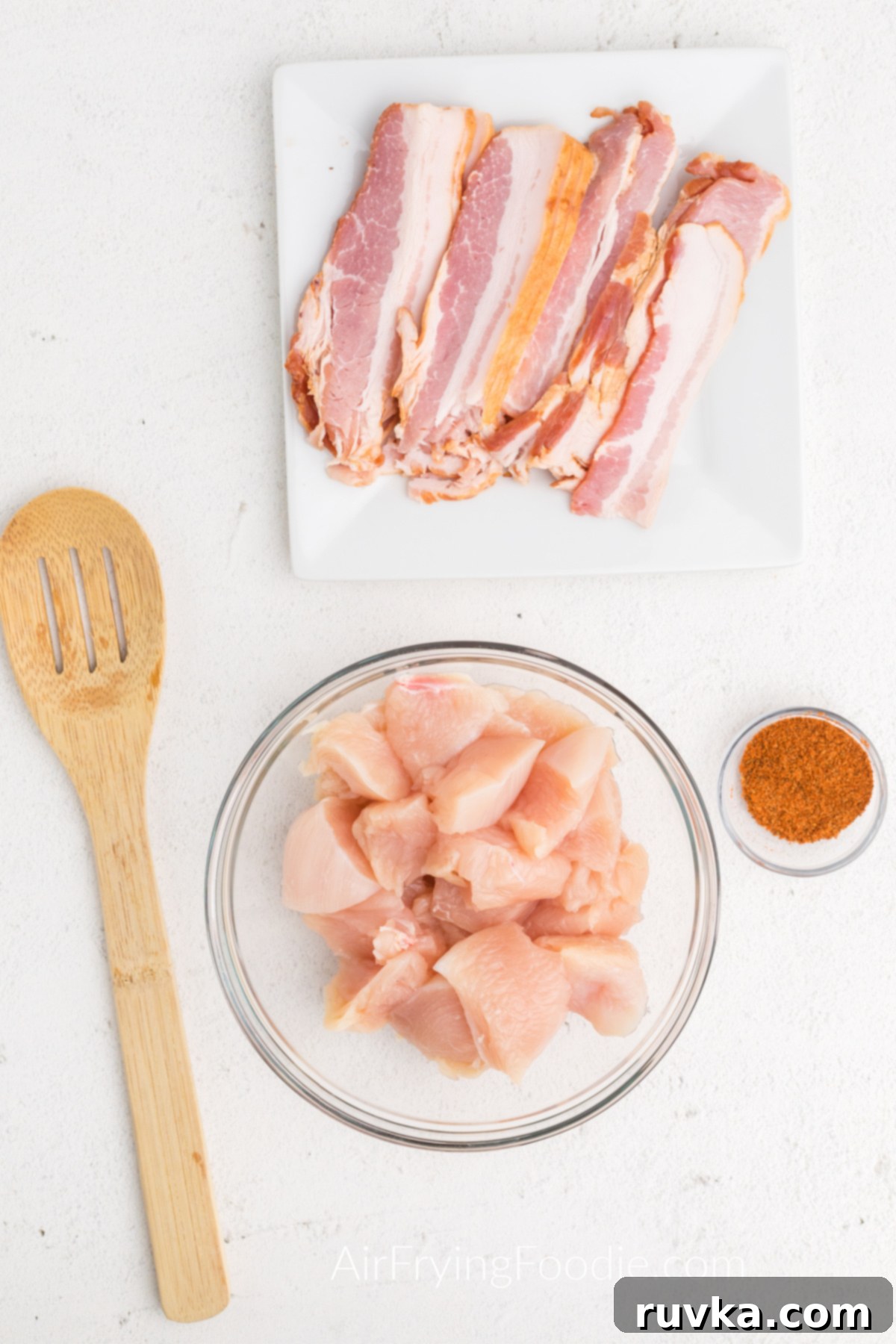 Perfectly Crispy Air Fryer Bacon Wrapped Chicken Bites 4 Bacon on a white plate, seasonings in a small container, and chicken pieces in a bowl.