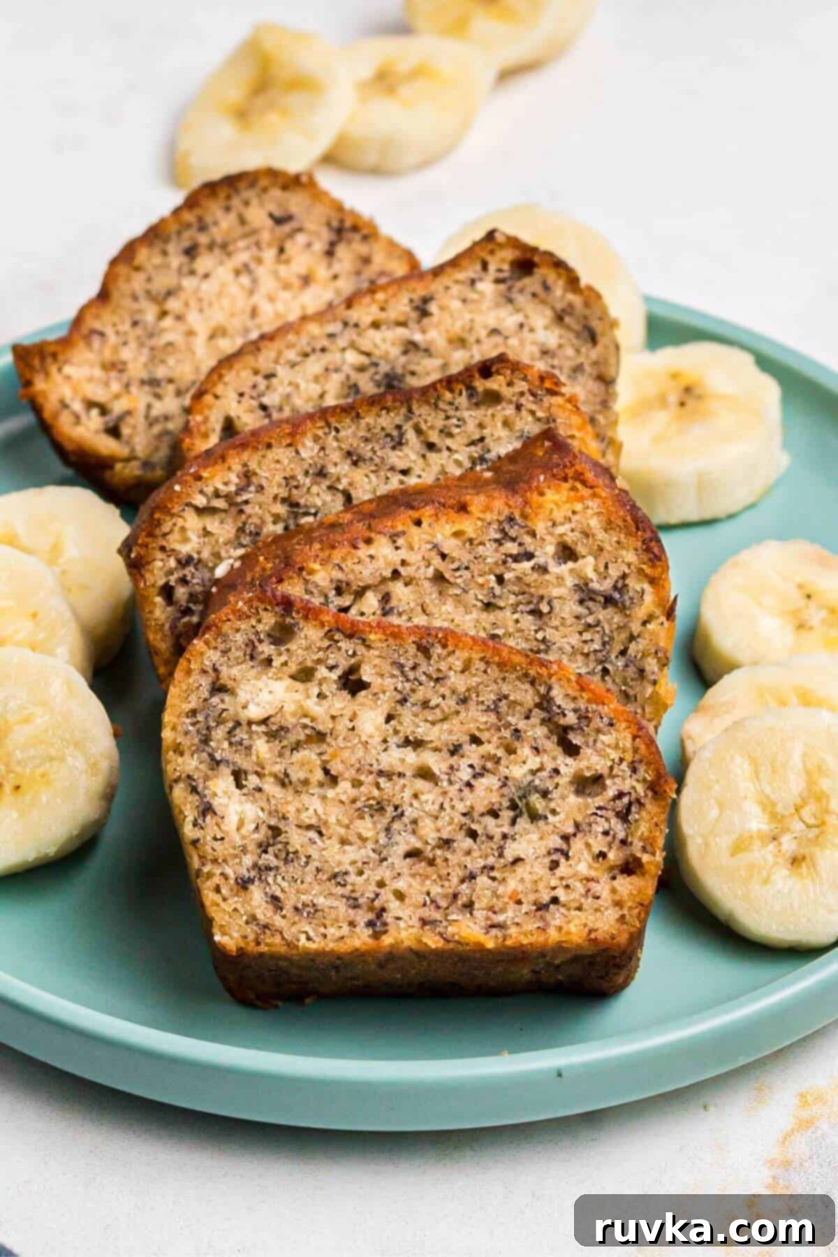 Golden brown baked banana bread sliced on a light green plate with slices of banana on the plate. 