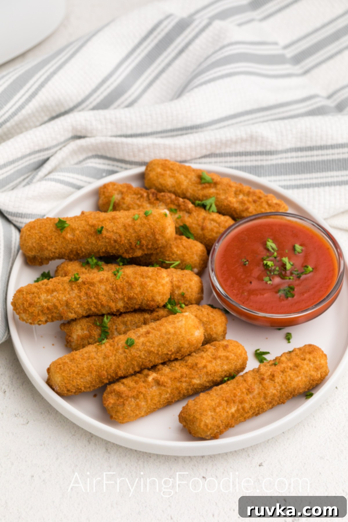 Crispy Air Fryer Mozzarella Sticks 2 Air Fryer Mozzarella Sticks on a white plate with dipping sauce and topped with fresh parsley.