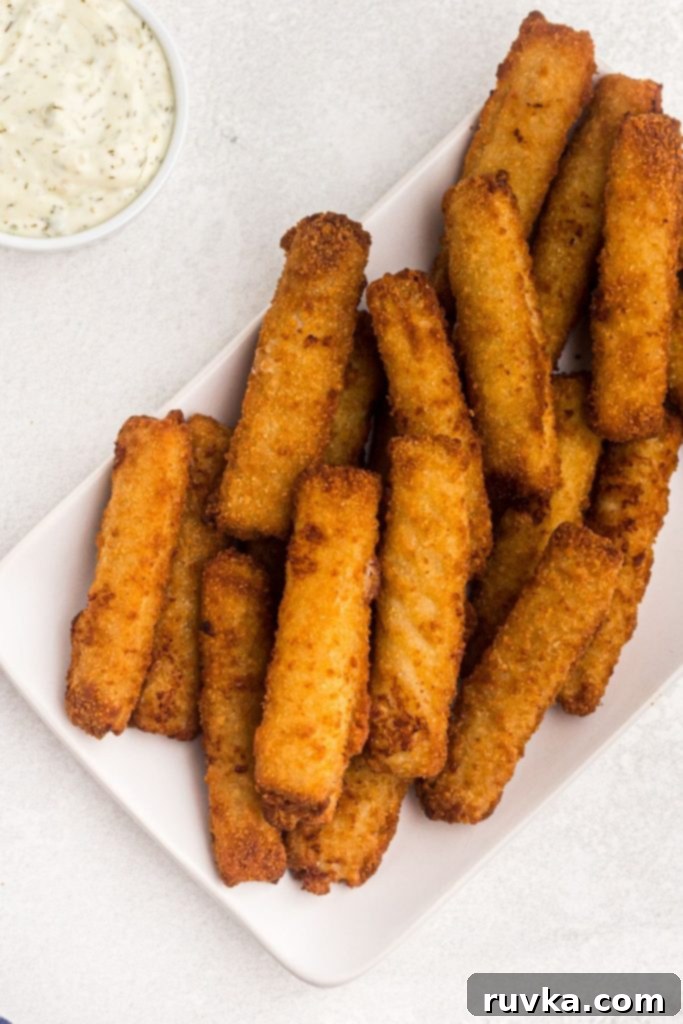 Crispy Air Fryer Fish Sticks 2 Golden fried fish sticks on a white marble table with a side dish of tartar sauce.