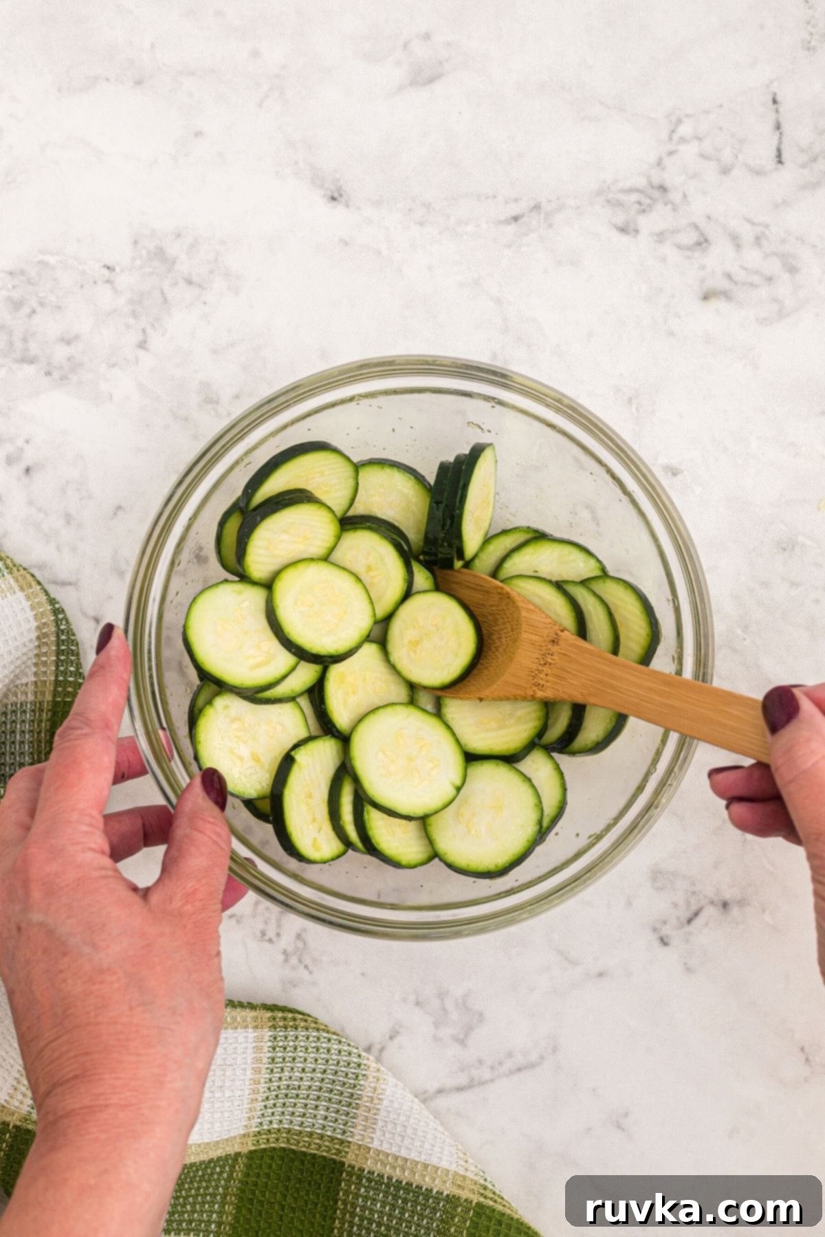 Golden Crispy Air Fryer Zucchini 4 Glass bowl with zucchini slices being tossed with olive oil and salt on a marble table.