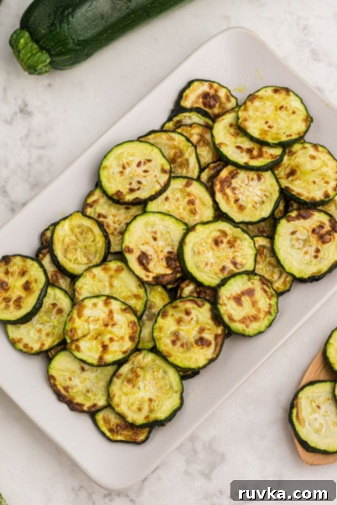 Golden Crispy Air Fryer Zucchini 8 Golden cooked zucchini slices on a white serving dish on a white marble table, ready to be enjoyed.