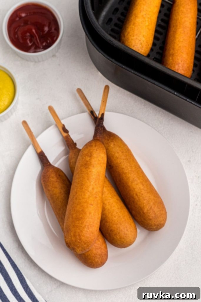 Golden Crispy Air Fryer Corn Dogs 2 Golden corn dogs on a white plate with ketchup and mustard on the table.