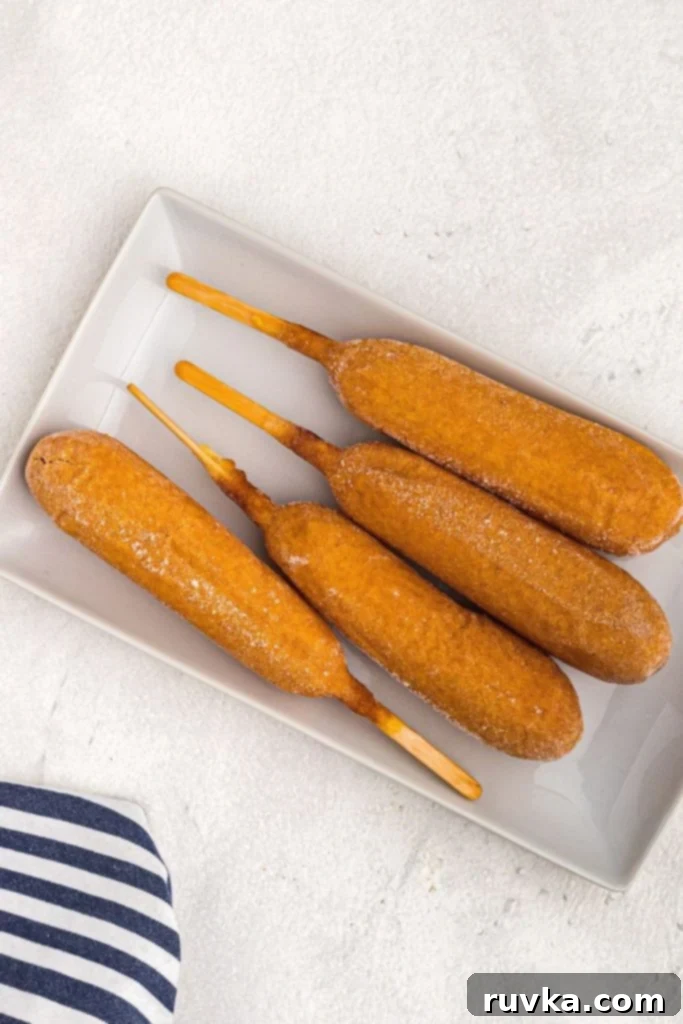 Golden Crispy Air Fryer Corn Dogs 3 Frozen corn dogs on a white plate on a marble table before being cooked in the air fryer.