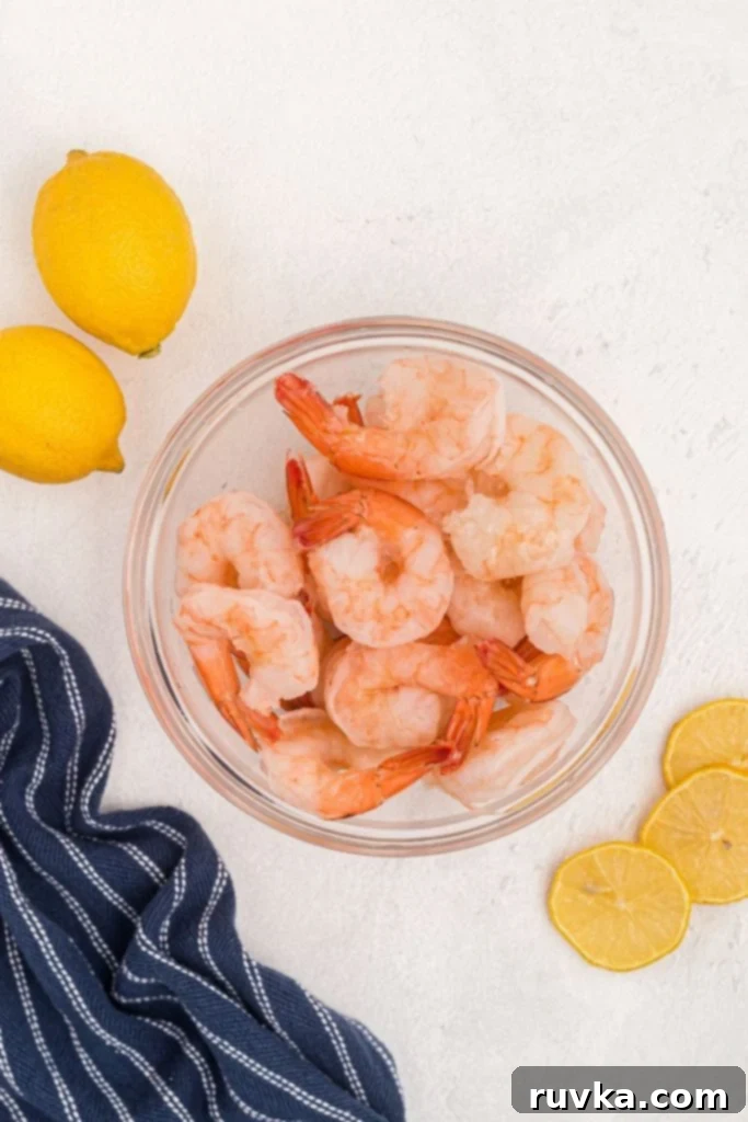 Easy Air Fryer Frozen Shrimp 3 Frozen shrimp in a clear glass bowl with lemon slices on a white marble table, ready for seasoning.