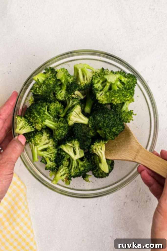 Crispy Air Fryer Broccoli 4 Uncooked broccoli in a large clear glass bowl being tossed with olive oil and seasonings.