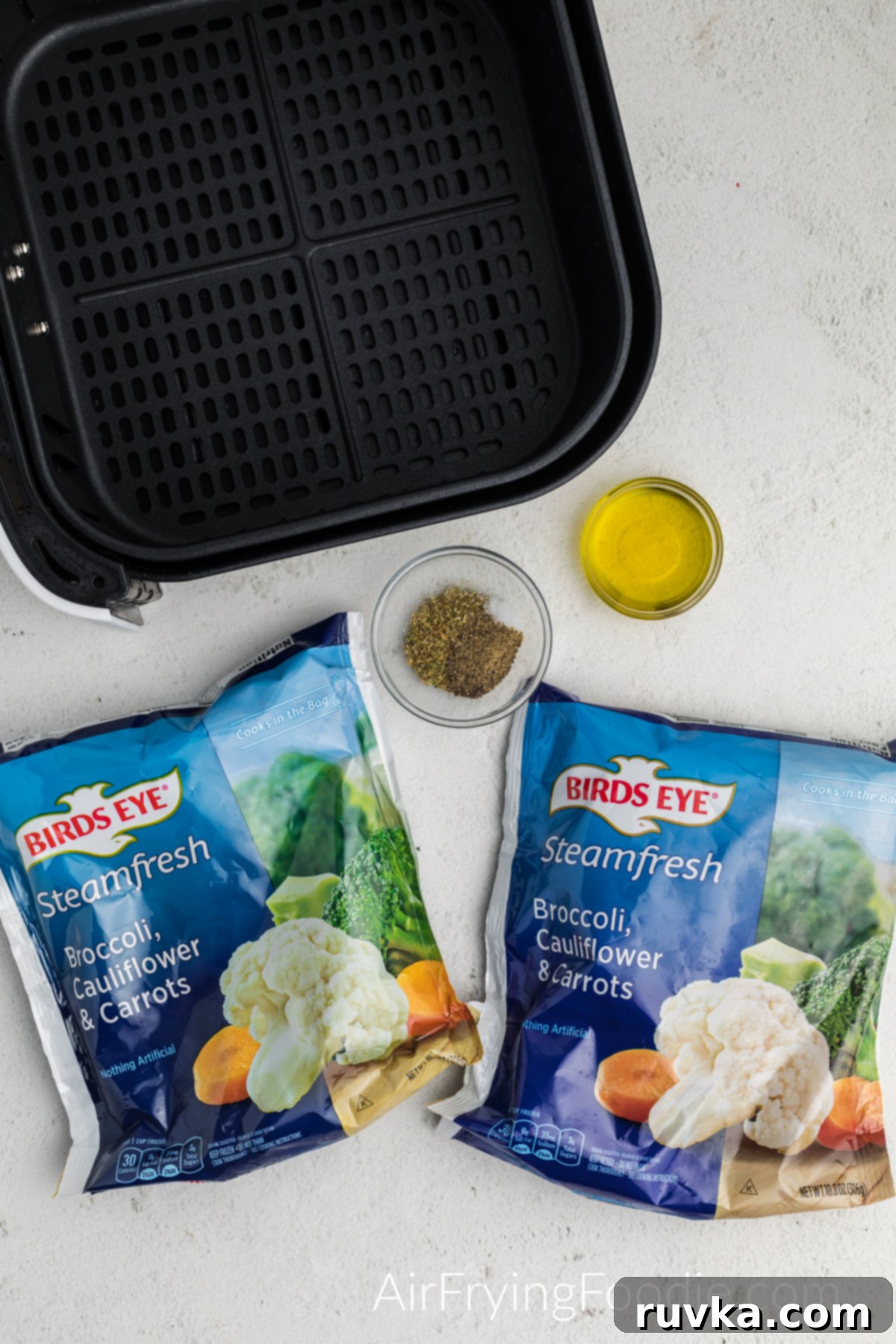 Crispy Air Fried Frozen Vegetables 3 Two bags of frozen mixed vegetables, a bottle of avocado oil, and various seasoning containers displayed with an air fryer basket on a white table, illustrating the simple ingredients.