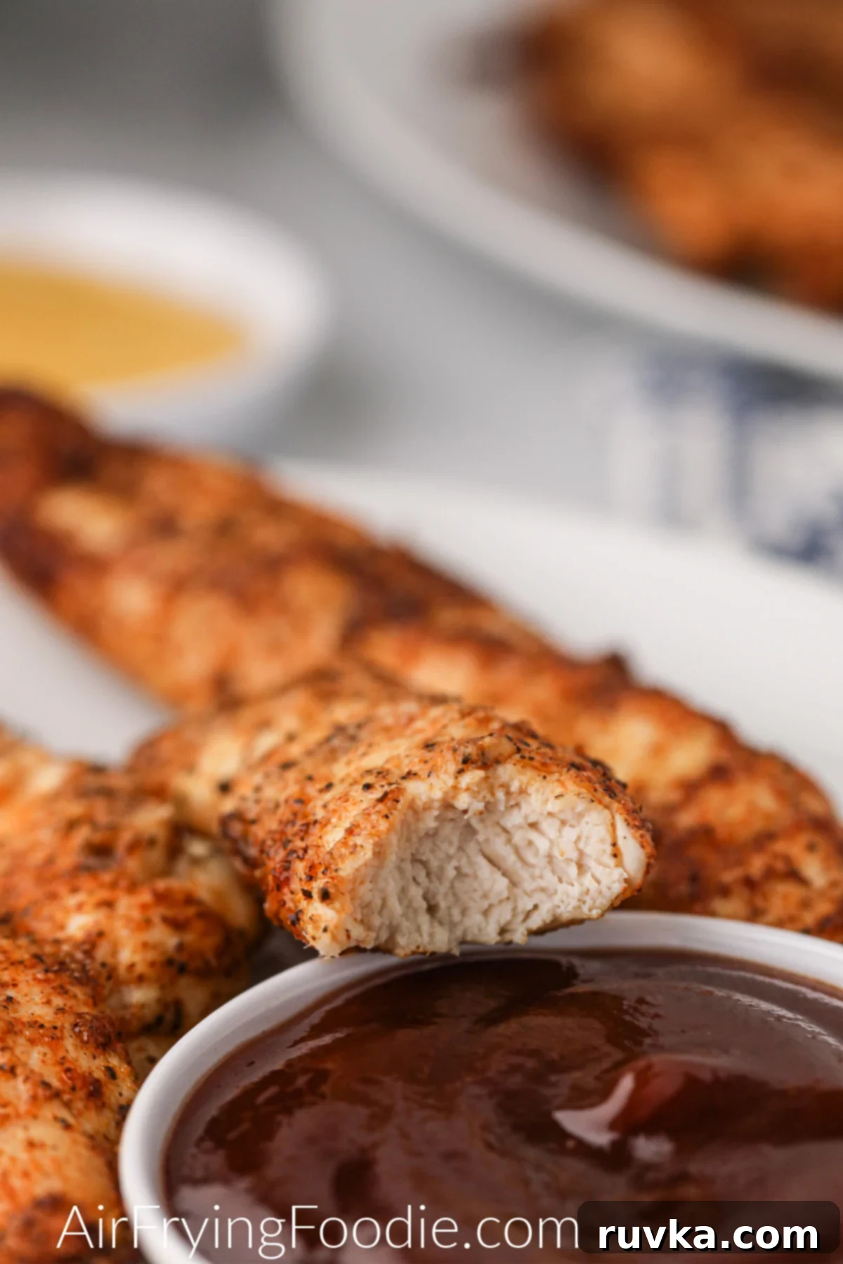 Naturally Crispy Air Fryer Chicken Tenders 6 Chicken tender with a bite taken out of it over a small bowl of BBQ sauce.