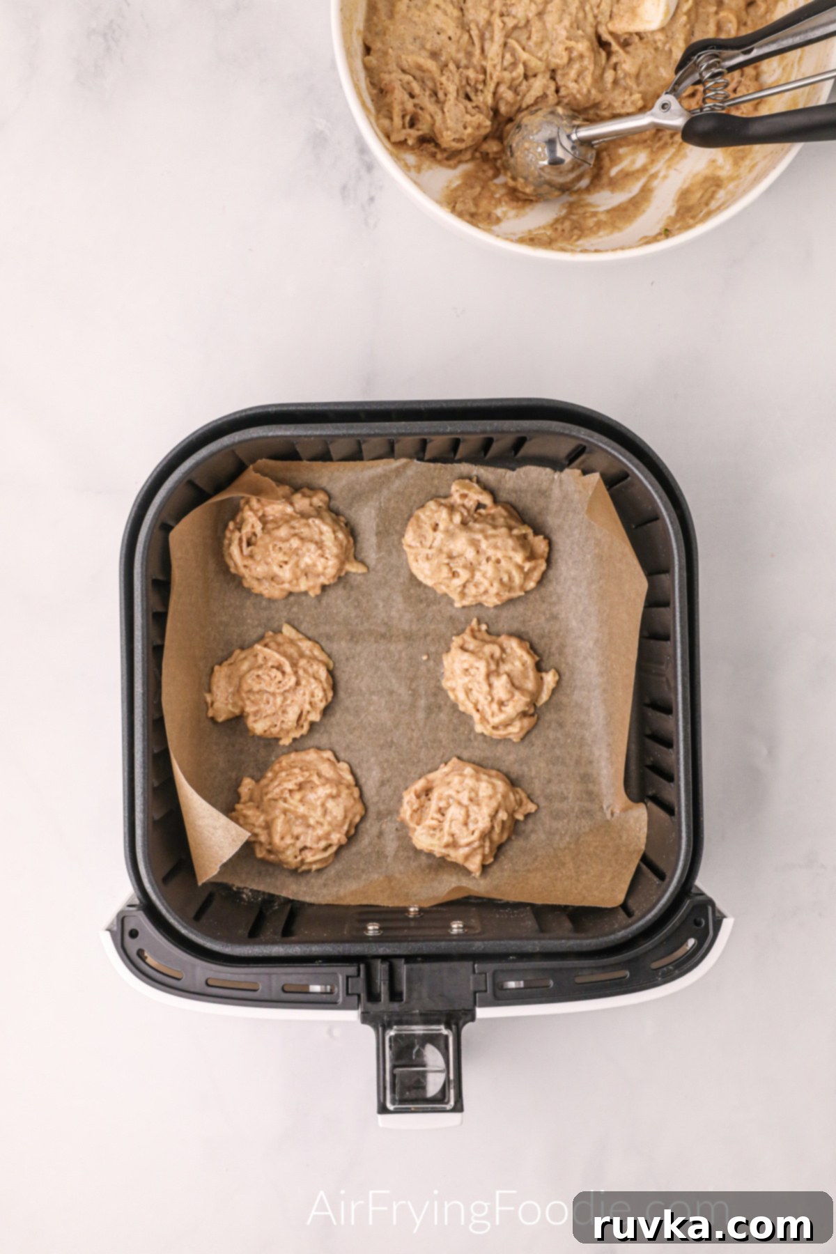 Easy Air Fryer Apple Fritters 6 Raw apple fritter dough, scooped in 2-tablespoon dollops, resting on parchment paper in an air fryer basket, ready for cooking.