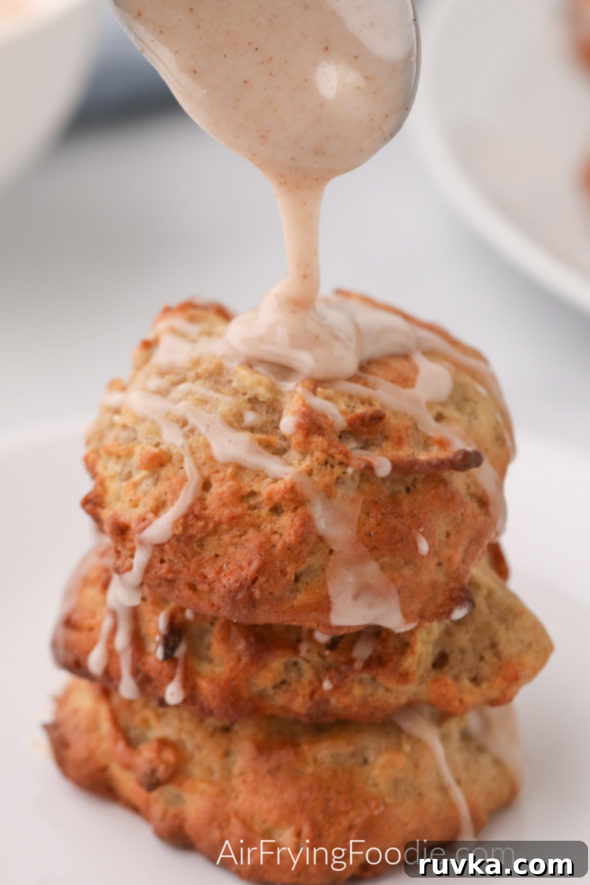Easy Air Fryer Apple Fritters 9 A stack of golden brown air fryer apple fritters on a white plate, with a spoon drizzling fresh glaze over the top, highlighting their deliciousness.